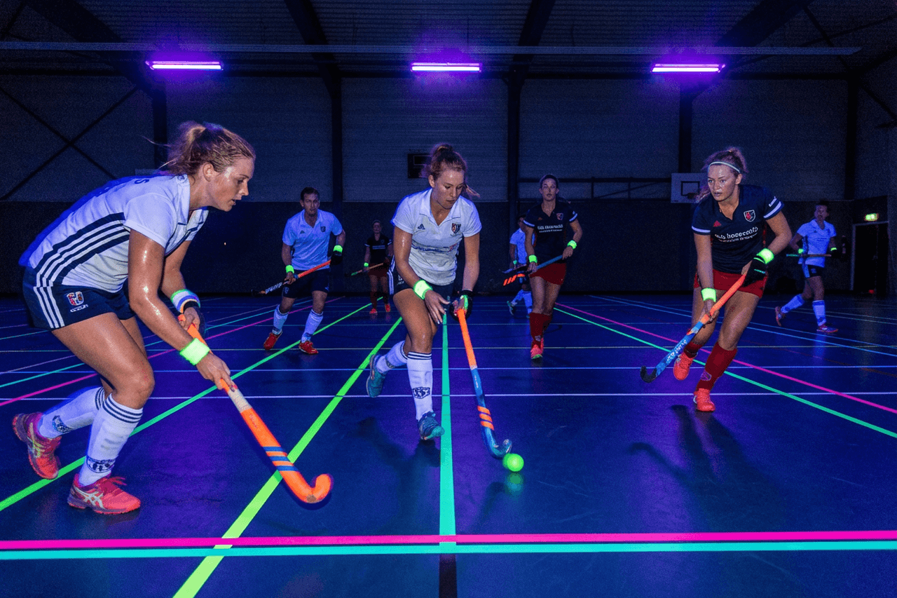 Glow in the dark zaalhockey in een sporthal met blacklight en neon lijnen.