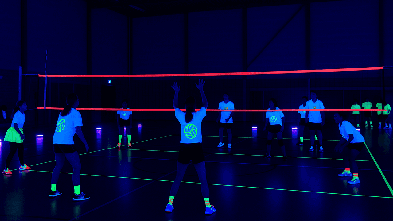 Blacklight volleybal in een sporthal met neonlijnen, UV-verlichting en spelers in fluorescerende kleding.